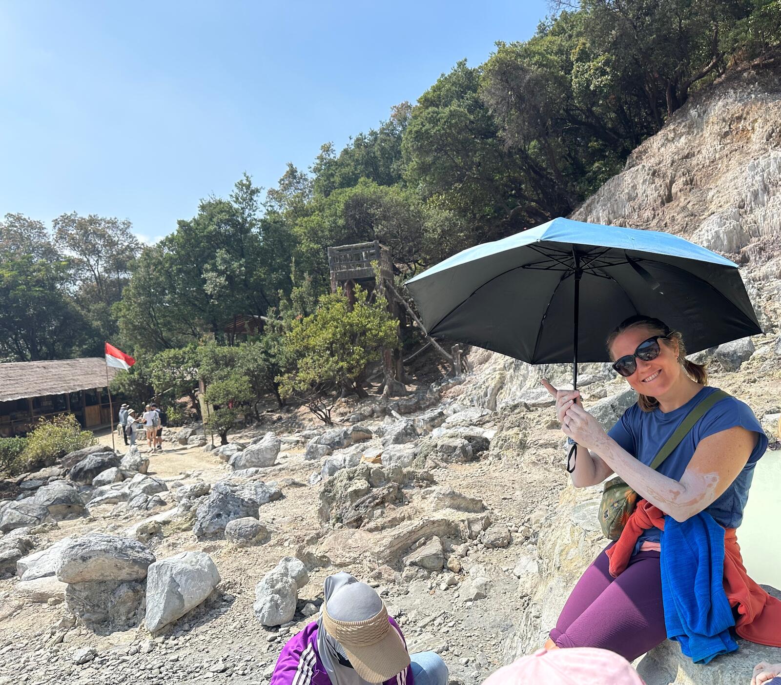 An image of Vikki sitting on the edge of a volcanic pool, with volcanic mud on her arm and a lady bent down massaging her legs with mud.