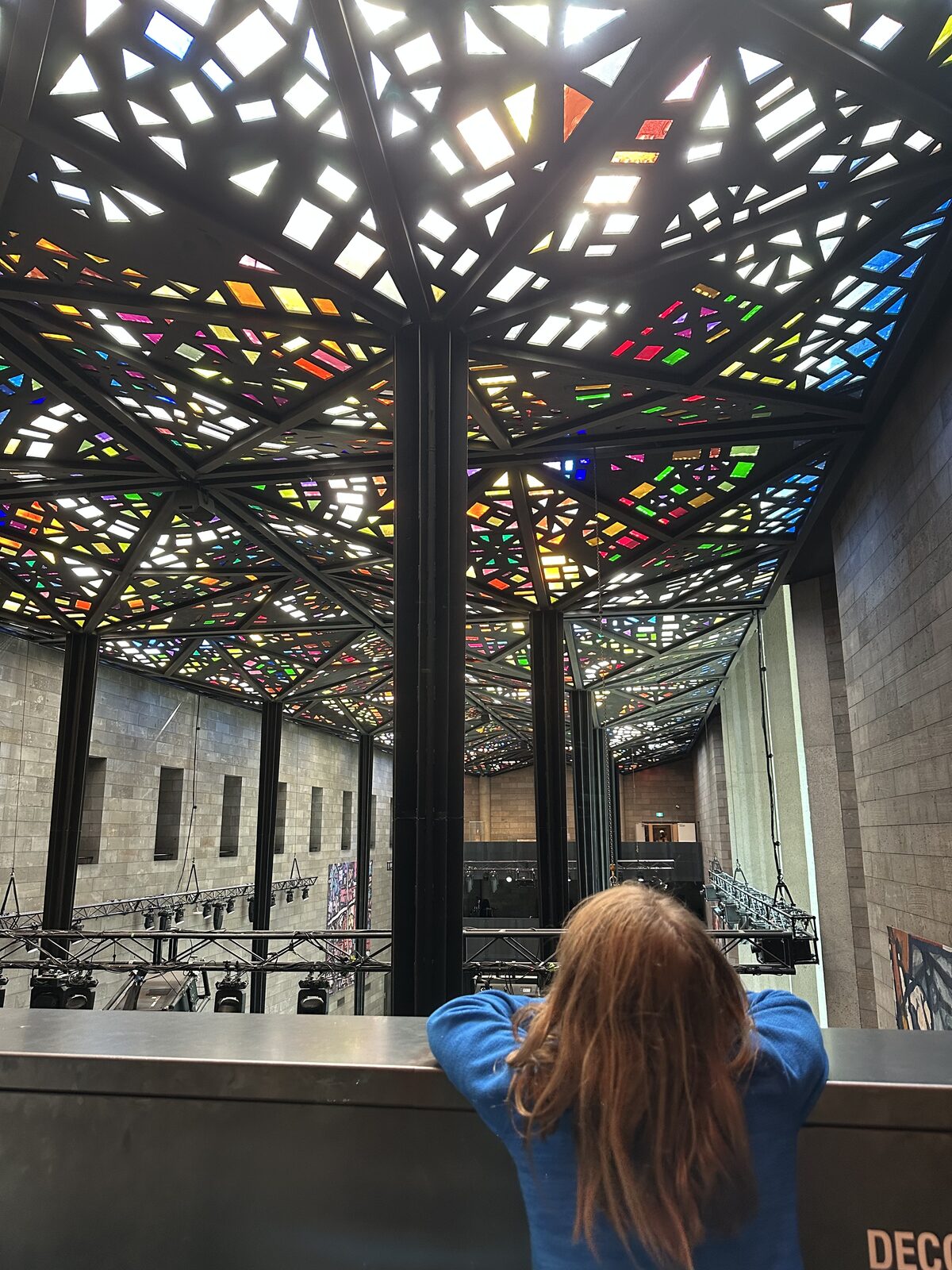 The back of a girl, looking up at a stained glass mosaic ceiling in the NGV