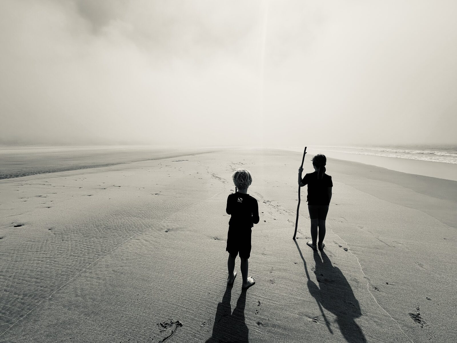 The silhouette of 2 children, standing looking out on a vast expanse of sandy beach in a foggy sky