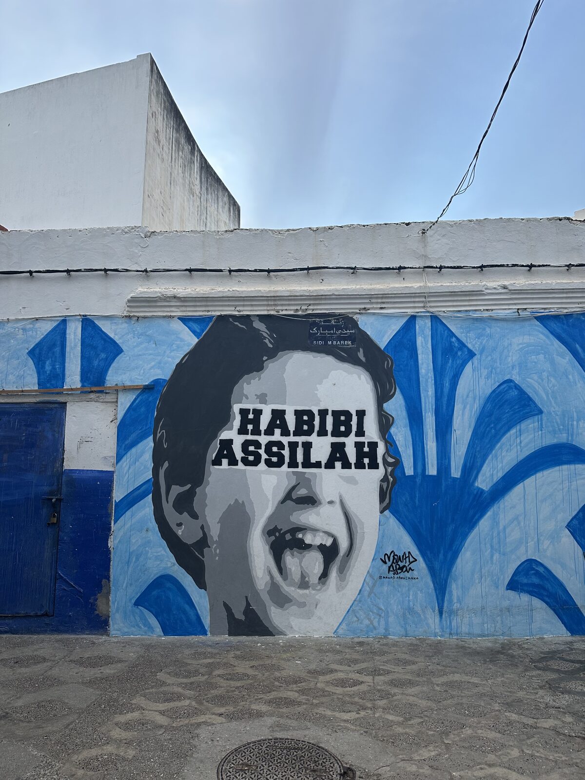 A photo of a wall of street art of a boys laughing face with the words “Habibi Assilah” on top