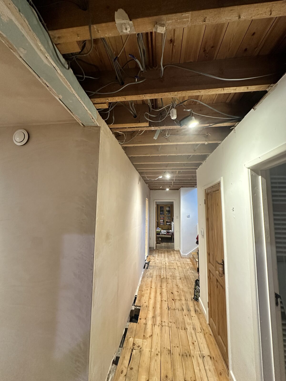 An internal hallway being renovated, with holes in the floorboards and the ceiling open with visible wires and new plasterboard