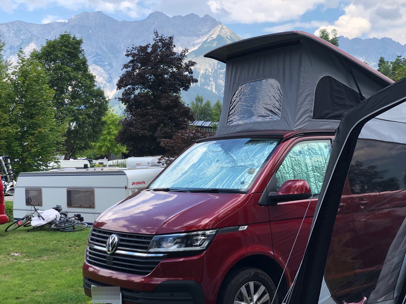 A red VW T6 campervan with pop-top up and the awning to the side in the foreground, with mountains rising up behind the campsite